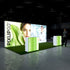 A side perspective of the Pixlip GO booth highlighting its seamless blend of nature and science. The illuminated panels display a mix of natural textures and a professional lab setting, while the counters maintain a minimalist green and white design. A visitor is seen approaching the booth.